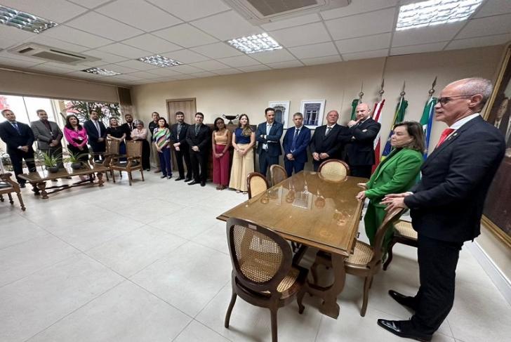 Fotografia em ambiente fechado. Sala de reunião com grupo de pessoas ao redor de uma mesa de madeira.
