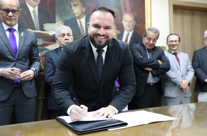 Fotografia em ambiente fechado, centralizado um homem branco, cabelos negros, sorrindo e usa terno azul. Ele assina documento.