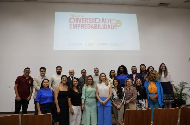 Fotografia em ambiente fechado de diversas pessoas em pé, lado a lado, formando uma fila mais acima e outra mais abaixo.