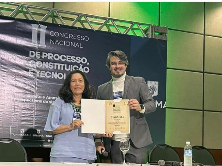 Fotografia em ambiente fechado que reúne duas pessoas em pé segurando um certificado. Fim da descrição.