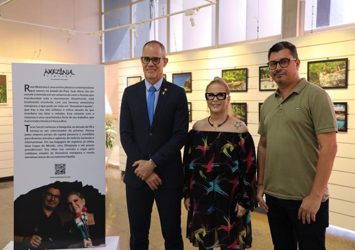 Fotografia em ambiente fechado composta por três pessoas em pé, sendo dois homens e uma mulher, lado a lado. Fim da descrição