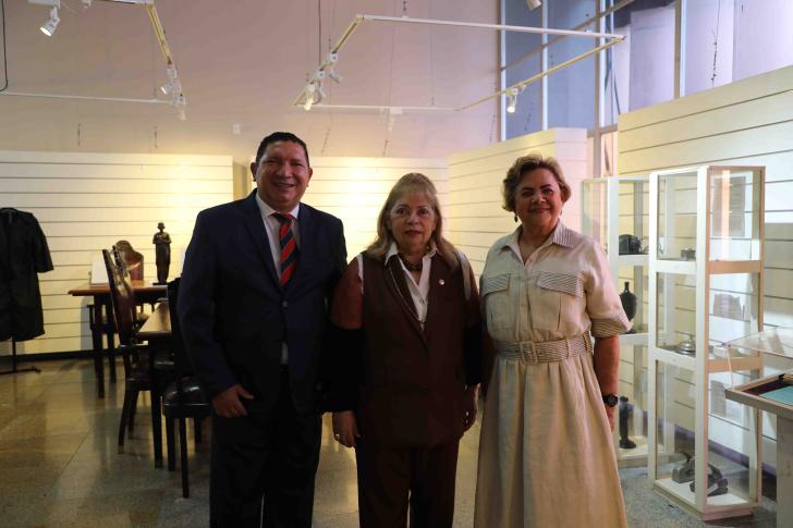 Foto colorida em ambiente fechado os presentes estão vestidos formalmente, com um homem de terno e mulheres com roupas sociais.
