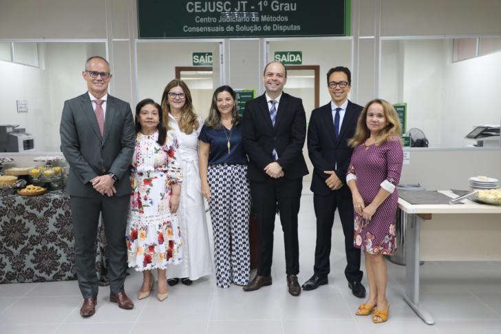 Foto colorida em ambiente fechado os presentes estão vestidos formalmente, com homens de terno e mulheres com roupas sociais.