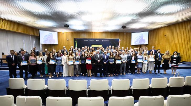 Foto colorida em ambiente fechado os presentes estão vestidos formalmente, com homens de terno e mulheres com roupas sociais.