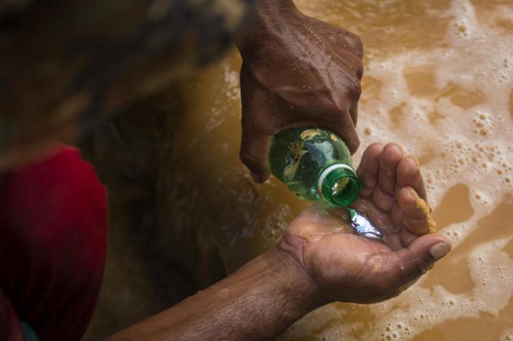 Close de mãos masculinas, de uma garrafa pequena verde é derramado mercúrio líquido na outra mão. A pessoa parece estar agachada
