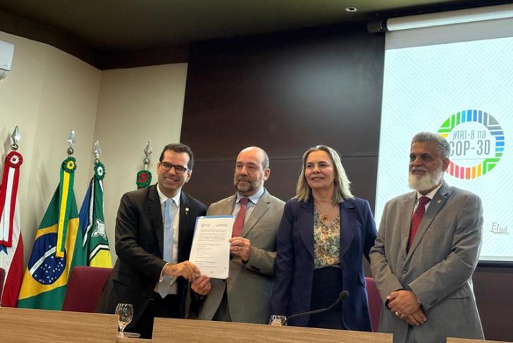 Foto colorida em ambiente fechado os presentes estão vestidos formalmente, com homens de terno e uma mulher com roupas sociais.