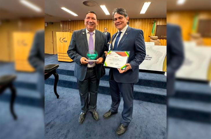Fotografia em ambiente fechado que reúne dois homens pardos, lado a lado, segurando premiações do CSJT