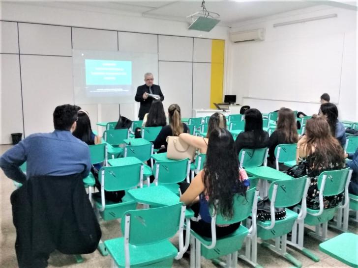 Na foto: Uma sala de aula com alunos sentados em cadeiras verdes e um palestrante em pé em frente à lousa.