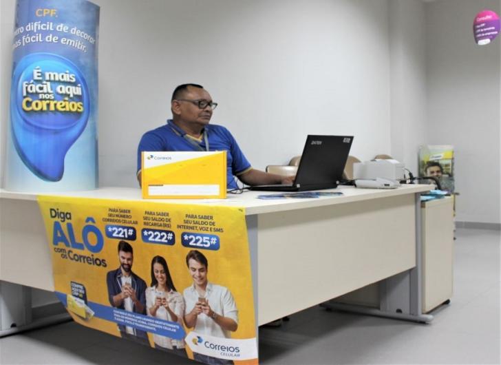 Na foto: Servidor dos Correios mexendo no notebook, na frente uma caixa de envio e um banner divulgando um serviço dos correios.