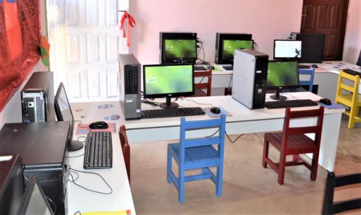 Na imagem: Laboratório com computadores doados sobre mesas e cadeiras coloridas.