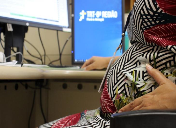 Na foto: Servidora grávida sentada na mesa de trabalho com a mão sobre a barriga e ao fundo monitor com a logo do TRT8.