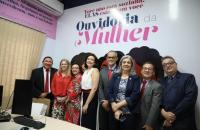 Oito pessoas lado a lado em uma sala com paredes adesivadas com o texto "Ouvidoria da Mulher"