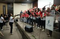 Fotografia em ambiente fechado. Um Coral formado por crianças e jovens que usam gorro de Papai Noel, blusa branco e casaco verme