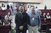 Fotografia em ambiente fechado. Ao centro, se encontram dois homens lado a lado.