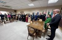 Fotografia em ambiente fechado. Sala de reunião com grupo de pessoas ao redor de uma mesa de madeira.