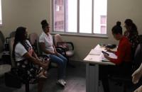 Fotografia em ambiente fechado com duas mulheres sentadas atrás de uma mesa e duas jovens em cadeiras em frente à elas.