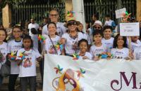 Foto em ambiente aberto, com mulheres, homens e crianças vestindo camisa branca e segurando um catavento colorido.