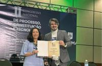 Fotografia em ambiente fechado que reúne duas pessoas em pé segurando um certificado. Fim da descrição. 