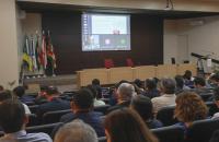  #ParaTodosVerem: Fotografia em ambiente fechado. Contendo dezenas de pessoas sentadas em um auditório. 