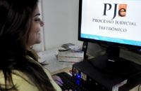 Fotografia colorida de uma mulher, sentada, vista de perfil, olhando para o computador e sorrindo. Ela é negra, cabelo escuro. 