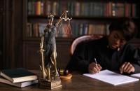 Foto colorida. No primeiro plano vemos a estátua que representa a Justiça. Ao fundo, uma magistrada negra parece assinar papéis,