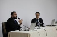 Fotografia em ambiente fechado de dois magistrados, durante uma palestra. Os dois homens são brancos e usam paletó preto.
