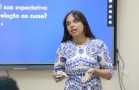 Fotografia da professora Olívia Rocha em sala de aula. Ela é uma mulher parda, de cabelos lisos, longos e escuros.