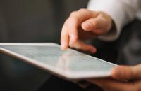 A imagem mostra a mão de um homem em destaque manuseando a tela de um tablet.