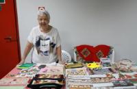 #ParaTodosVerem: Fotografia colorida em ambiente fechado, na foto está presente uma senhora sorrindo atrás de sua mesa com seus 
