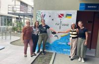 Fotografia em ambiente aberto. Na foto está presente 5 mulheres, todas estão sorrindo na frente de uma parede que acabou de ser 