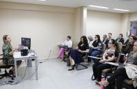 Foto colorida mostra momento de aula de precedentes. A professora está sentada à mesa e fala aos alunos.
