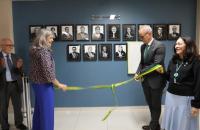 Fotografia em ambiente fechado que reúne duas mulheres e um homem, descerrando uma faixa de inauguração de exposição.