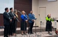 Fotografia em ambiente fechado que reúne um grupo musical formado por homens e mulheres e uma mulher branca em destaque. 