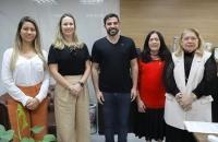 Descrição de imagem: Fotografia em ambiente fechado, nela, estão cinco pessoas, quatro mulheres e um homem. Todos estão de pé e 