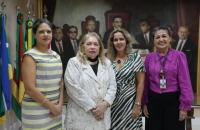 Descrição de imagem: Fotografia em ambiente fechado, nela, estão quatro mulheres de pé e sorrindo para a foto. 