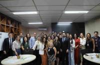 Foto colorida em ambiente fechado os presentes estão vestidos formalmente, com homens de terno e mulheres com roupas sociais.