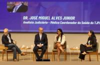 Descrição de imagem: Captura de tela contendo quatro pessoas sentadas em um palco enquanto seguram microfones