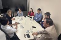 #ParaTodosVerem: Fotografia colorida em ambiente fechado. A foto mostra homens e mulheres sentados em reunião