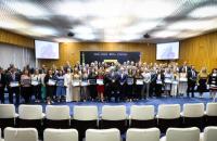 Foto colorida em ambiente fechado os presentes estão vestidos formalmente, com homens de terno e mulheres com roupas sociais.