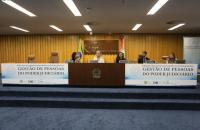 Foto mostra, em primeiro plano, mesa do evento com palestrantes e ao fundo logomarca do TST.