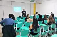 Na foto: Uma sala de aula com alunos sentados em cadeiras verdes e um palestrante em pé em frente à lousa.
