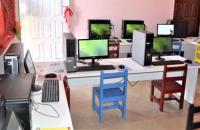Na imagem: Laboratório com computadores doados sobre mesas e cadeiras coloridas.