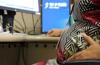 Na foto: Servidora grávida sentada na mesa de trabalho com a mão sobre a barriga e ao fundo monitor com a logo do TRT8.