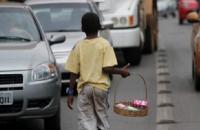 Foto mostra menino segurando cesta com coisas a venda, transitando entre veículos, em avenida.