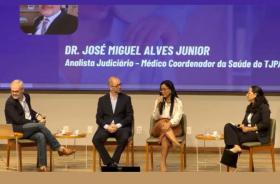 Descrição de imagem: Captura de tela contendo quatro pessoas sentadas em um palco enquanto seguram microfones